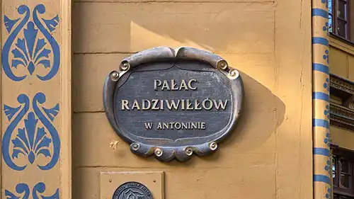 Ozdobna metalowa tablica z napisem Pałac Radziwiłłów w Antoninie, przymocowana do drewnianej ściany pałacu.