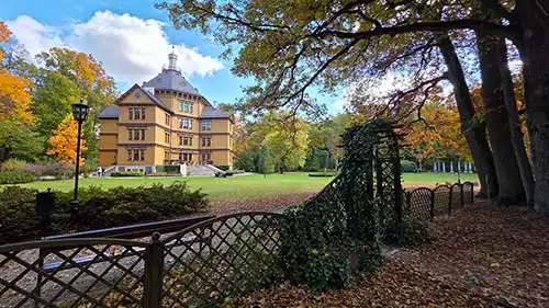 Pałac Myśliwski Radziwiłłów z Antonina widoczny zza drewnianego ogrodzenia w parku, po lewej latarnia i jesienne drzewa.