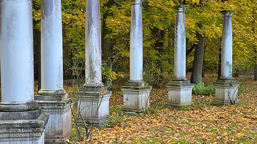 Rząd sześciu zabytkowych, klasycystycznych kolumn na postumentach w jesiennym parku krajobrazowym Pałacu Radziwiłłów.
