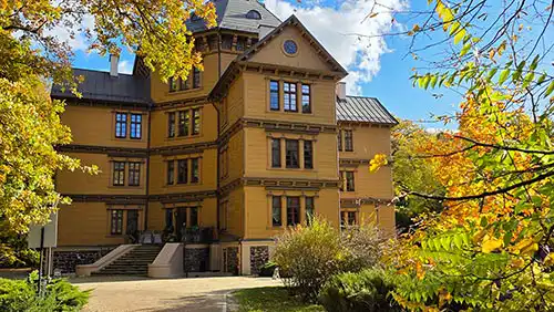 Drewniany Pałac Radziwiłłów w Antoninie, widok na główną fasadę otoczoną jesiennymi liśćmi i błękitnym niebem.