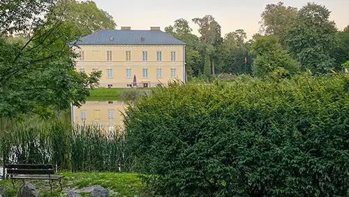 Pałac w Dobrzycy na tle zieleni parkowej z odbiciem w wodzie i ławką na pierwszym planie.