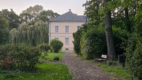 Ścieżka parkowa z ławką prowadząca do bocznej ściany Pałacu w Dobrzycy otoczonej bujną zielenią.