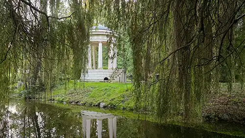 Klasycystyczna rotunda Monopter w Dobrzycy widziana przez wierzby płaczące i odbita w stawie.