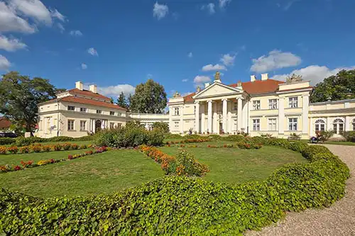 Szeroki widok na klasycystyczny Pałac Śmiełów i otaczający go park z wypielęgnowanym trawnikiem oraz żywopłotami.