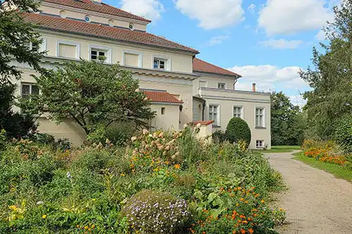 Boczne skrzydło Pałacu Śmiełów otoczone ścieżką parkową oraz naturalistycznym ogrodem bylinowym i krzewami.