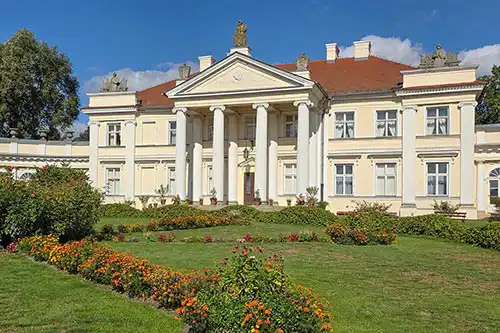 Zbliżenie na centralny korpus Pałacu w Śmiełowie, z portykiem jońskim i rzeźbą na dachu, otoczony barwnymi rabatami kwiatowymi.