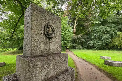 Pomnik Adama Mickiewicza z medalionem w parku Pałacu Śmiełów, widoczna ścieżka spacerowa.