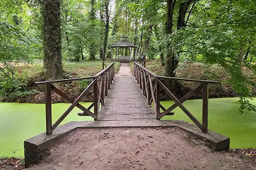 Drewniany mostek w parku Pałacu Śmiełów, prowadzący nad ciekiem wodnym do parkowej altany i zielonej alei.