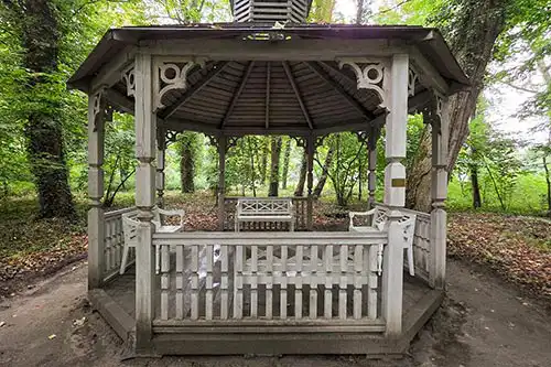 Ośmiokątna drewniana altana parkowa z ozdobnymi detalami i ławkami w gęsto zalesionym parku Pałacu Śmiełów.