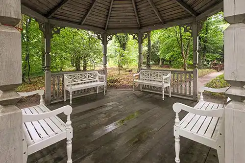 Wnętrze ośmiokątnej altany parkowej Pałacu Śmiełów: drewniana podłoga i białe ławki z widokiem na leśne otoczenie.