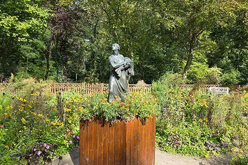 Rzeźba Zosi z Pana Tadeusza na drewnianym postumencie, otoczona ogrodem bylinowym przy Pałacu Śmiełów.