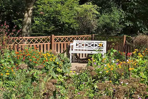 Biała drewniana ławka w stylizowanym Ogrodzie Zosi Pałacu Śmiełów, otoczona kolorowymi bylinami i ażurowym płotem.