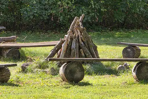 Miejsce na ognisko w parku Pałacu Śmiełów: ułożone w stożek drewno opałowe otoczone prostymi ławkami z pni.